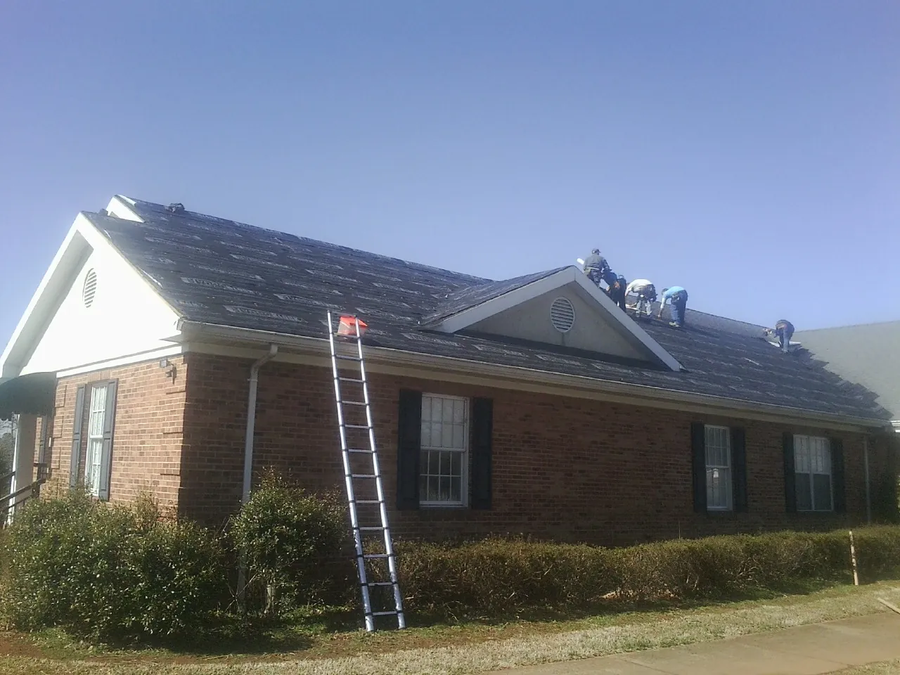 Crew tearing shingles off a brick ranch roof with a ladder set — Emergency Roof Repair work by our crew in Fosters