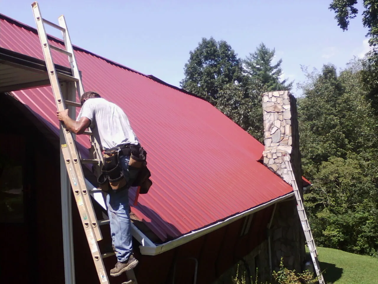Roofer on a ladder beside a red metal roof and stone chimney — Soffit & Fascia Repair by Roofing Brothers in Fosters